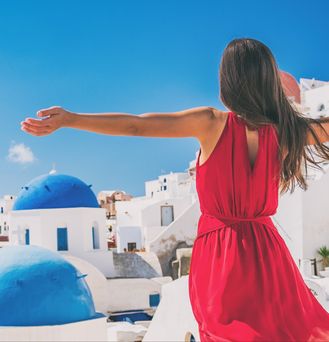 Eine brünette Frau in rotem Sommerkleid steht mit weit auseinandergebreiteten Armen vor Santorin. Ein Meer aus weißen Häusern mit blauen Dächern erstreckt sich vor dem blauen Meer.