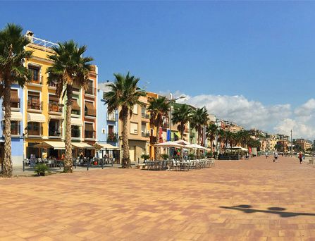 An einer bepflasterten Strandpromenade stehen Palmen, dahinter Restaurants mit Außenplätzen und Markisen und bunten Häuserfassaden.