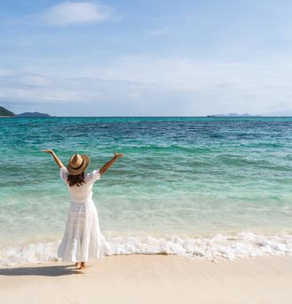 Eine Frau mit weißem Sommerkleid und Sonnenhut steht vor dem Meer und reißt die Arme freudig in die Luft.