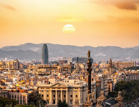 Die Skyline einer spanischen Stadt erstreckt sich in gold, gelbem Licht. Im Hintergrund sind dunkle, große Berge und darüber die runde, untergehende Sonne.