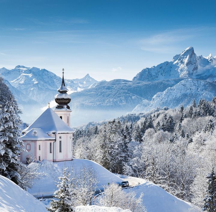 Eine kleine, weiß-rosa-farbene Kapelle ragt aus einer winterlichen Landschaft mit schneebedeckten Tannen und Berggipfeln hervor.