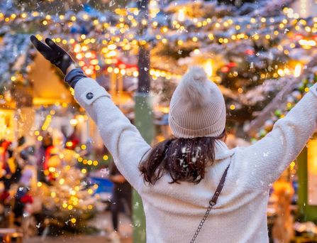 Eine Frau steht dem Betrachtenden mit dem Rücken zugewendet und armen in der Luft auf einem Weihnachtsmarkt. Sie trägt Mütze und Handschuhe und im Hintergrund sieht man viele gold-glitzernde Lichter.