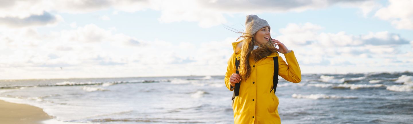 Eine junge Frau mit gelber Regenjacke und grauer Mütze steht am menschenleeren Strand. Der Wind weht ihr durch die Haare und sie blickt aufs Meer.