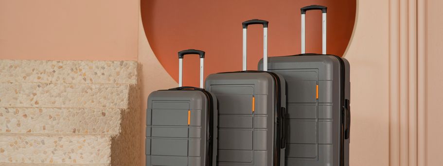 3 graue Rollkoffer stehen der Größe nach geordnet vor einer orangenen Wand.