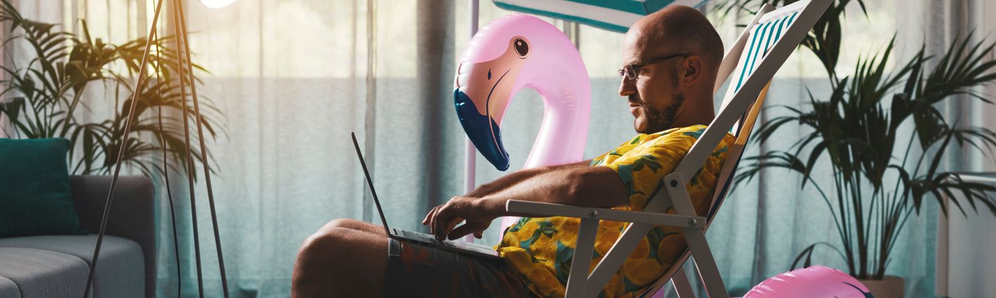 Ein Mann sitzt im Hawaii Hemd im Liegestuhl mit Laptop auf dem Stuhl in einem Raum. Neben ihm ein pinker Gummi-Flamingo.