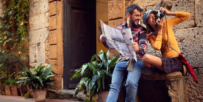 Reisetrends 2026: Ein junge Paar sitzt auf einer Bank in einer mediterranen Gasse mit Stadtplan und Kamera
