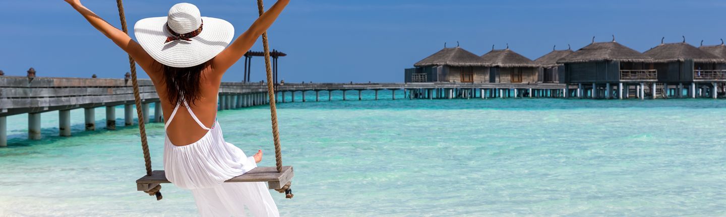 Malediven Urlaub: Eine Frau mit weiße Sommerkleid und Hut sitzt auf einer Schaukel am Strand.