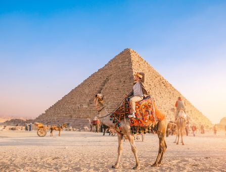 Ein Mann mit Sonnenhut sitzt auf einem bunt geschmückten Kamel. Im Hintergrund weitere Kamele und Menschen und eine Pyramide.