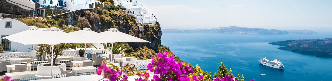An einem Abhang mit mediterranen Pflanzen befindet sich eine Art Plateau auf welchem weiße Stühle und Sonnenschirme stehen. Unten in einer Schlucht gleitet ein weißes Kreuzfahrtschiff auf dem spiegelglatten Meer.