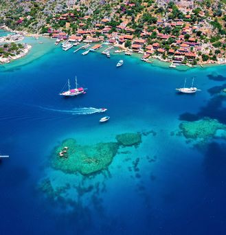 Eine Luftaufnahme einer Bucht mit tiefblauem Meer, türkisen Unterwasserinseln und einigen (Segel-)Booten.