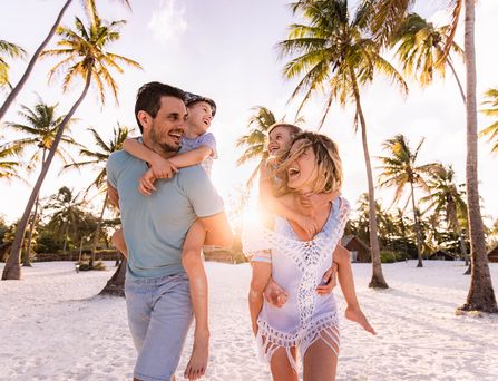 Eine junge vierköpfige Familie läuft lachend an einem Strand entlang. Der Vater hat den Sohn auf dem Rücken und die Mutter die Tochter. Alle tragen Sommerkleidung und lachen sich an.
