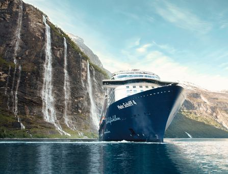 Das blau-weiße Kreuzfahrtschiff "Mein Schiff 4" gleitet durch das dunkle Meer entlang eines steilen Felsens mit Wasserfall.