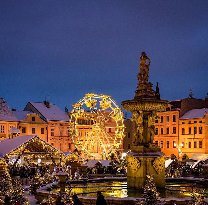 In der Abenddämmerung befindet sich ein Marktplatz umgeben von hübschen, Altbauten. Auf dem Platz ist ein beleuchtetes Riesenrad, Brunnen und weihnachtliche Hütten.