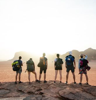 Eine Gruppe aus sechs jungen Menschen mit Rucksäcken und Outdoorausrüstung steht auf einem Felsen und blickt in eine Felslandschaft.