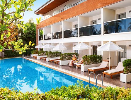 Um einen großen türkisen Pool stehen weiße mehrstöckige Häuser mit Palmen und Sonnenliegen davor.