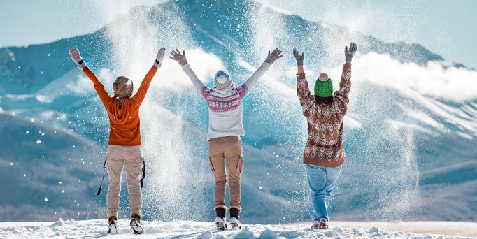 3 Menschen tragen Winterkleidung und werfen pulvrigen Schnee in die Luft. Im Hintergrund sieht man eine schneebedeckte Winterlandschaft