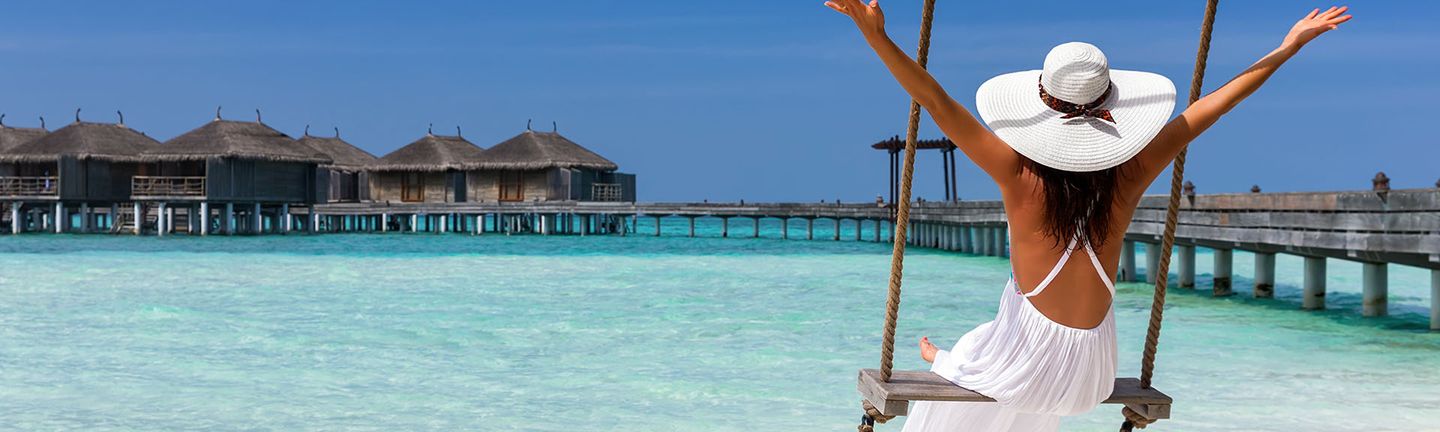 Malediven: Eine Frau in weißem Kleid und mit Sonnenhut schaukelt auf einer Holzschaukel und reißt die Arme in die Luft. Sie blickt auf den glasklaren türkisen Ozean mit einem Holzsteg und Wasserbungalows.