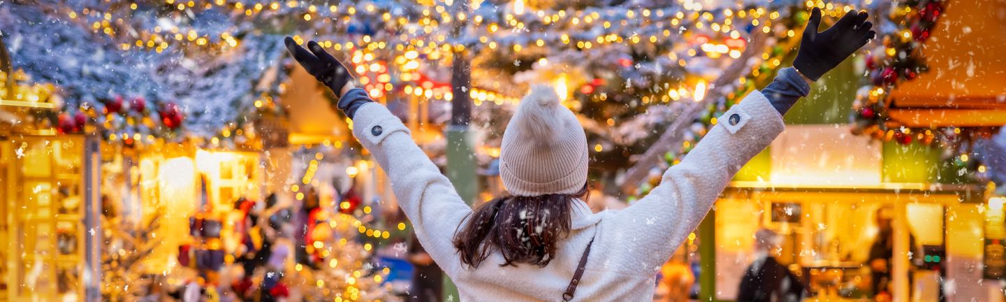 Eine Frau steht dem Betrachtenden mit dem Rücken zugewendet und armen in der Luft auf einem Weihnachtsmarkt. Sie trägt Mütze und Handschuhe und im Hintergrund sieht man viele gold-glitzernde Lichter.