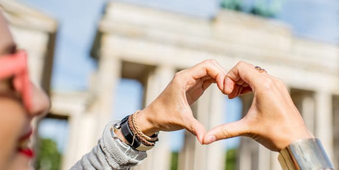 Eine Frau formt mit ihren Händen ein Herz und im Hintergrund ist unscharf das Brandenburger Tor zu sehen.