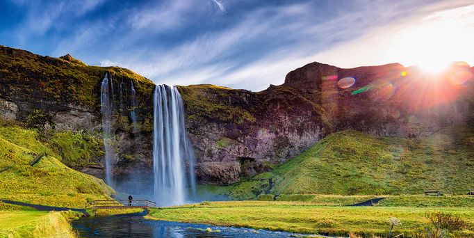 Eine grüne, felsige Landschaft mit untergehender Sonne und Wasserfall.