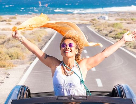 Eine sommerlich, hippimäßig gekleidete Frau sitzt in einem Cabrio mit offenem Dach. Sie steht im Auto und reist die Arme lachend nach oben und ein orangenes Tuch weht im Wind. Im Hintergrund das wilde Meer.
