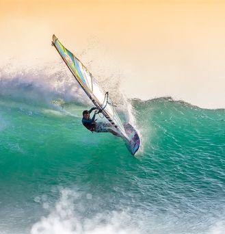 Ein Windsurfer steht fast senkrecht eine grün-türkise Welle nehmend auf dem Wasser.