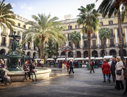 Belebter Platz mit mehreren Palmen, einem Brunnen und umliegenden historischen Gebäuden unter blauem Himmel.