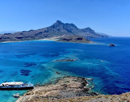 Eine Frohnenaufnahme von einem zerklüfteten Bucht mit einem angelegten Boot. Das Meer strahl tiefblau und im Hintergrund ragt ein Berg hervor.