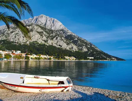 Ein weißes, kleines Motorboot liegt am Kiesstrand unter einer Palme und im Hintergrund ragt ein gigantischer grau, begrünter Berg empor