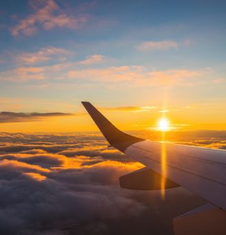 Der Blick auf einem Flugzeugfenster zeigt den linken Flügel. Das Flugzeug befindet sich über den Wolken und die Sonne fängt gerade an unter zu gehen.