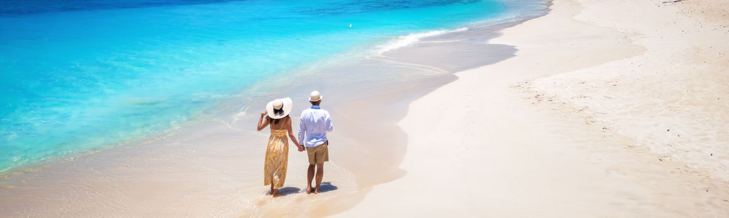 Ein Paar mit Sommerkleidung läuft Hand-in-Hand an einem weißen Sandstrand mit kristallklarem türkisen Wasser entlang.