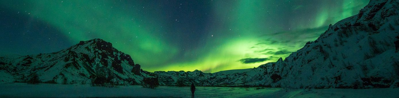 Der dunkel Nachthimmel wird von grünen Polarlichtern durchzogen. Eine Person steht, mit dem Rücken zugewandt und Wanderstöcken im weißen Schnee. Dahinter schneebedeckte Berge.