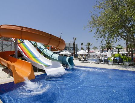Um einen großen türkisen Pool stehen weiße mehrstöckige Häuser mit Palmen und Sonnenliegen davor.