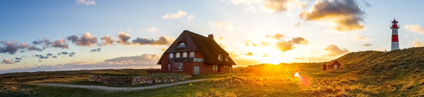 Ein großes rotes Haus mit Strohdach steht einsam in einer weiten, grünen Landschaft. Der Himmel ist von der untergehenden Sonne rosa-gold gefärbt und ganz rechts steht ein rot-weißer Leuchtturm.