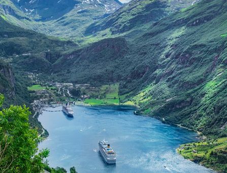 Inmitten eines norwegischen, saftgrünen Fjords befinden sich 2 Hochseekreuzfahrtschiffe. EIns liegt am Hafen an und ein weiteres bewegt sich von ihm weg.