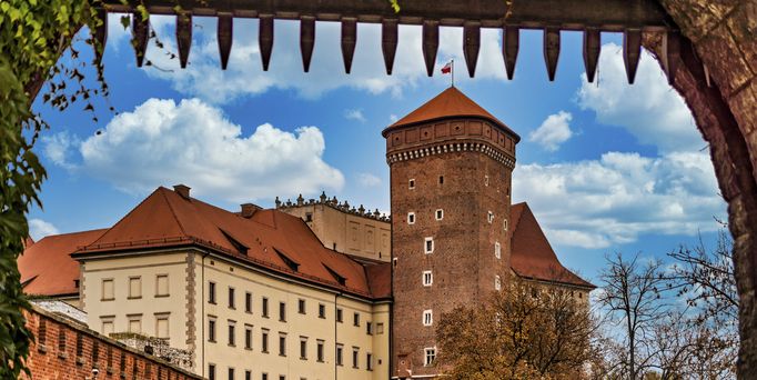 Ein mittelalterlicher Torbogen mit spitzem Gitter ragt am oberen Bildrand hervor. Im Hintergrund stehen gelbe Häuser mit orangenen Dächern und einem mittelalterlichen Burgturm.