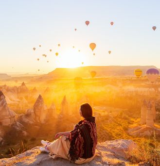 Eine Frau sitzt an einem Felsabsprung und blickt auf die Schlucht von Kappadokien. DIe Sonne geht gerade unter und taucht das gesamte Bild in ein orangenes Licht. Unzählige Heißluftballons schweben in der Luft.