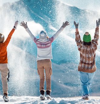 3 Menschen tragen Winterkleidung und werfen pulvrigen Schnee in die Luft. Im Hintergrund sieht man eine schneebedeckte Winterlandschaft