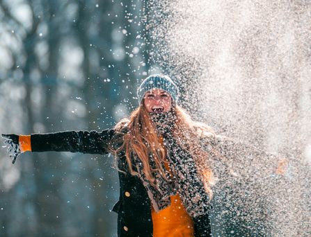 Eine junge blonde Frau mit Mütze und Handschuhen breitet lachend die Arme aus und wirbelt pulvrigen Schnee hoch.