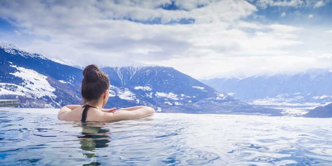 Eine Frau mit braunem Dutt lehnt am Rand eines Infinitypools mit Blick auf die verschneiten Bergspitzen.