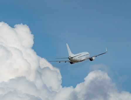 Ein weißes Flugzeug fliegt schräg durch den blauen Himmel über weißen puffrigen Wolken.