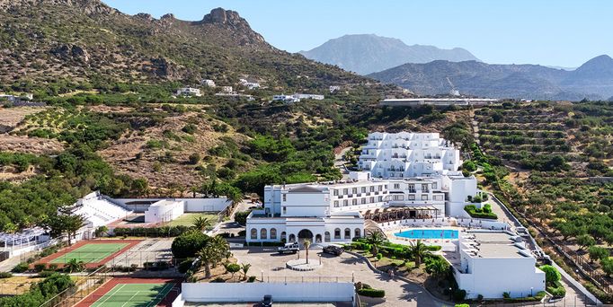 In einer begrünten Berglandschaft befinden sich eine weiße Hotelanlage mit Pool und mehreren Sportplätzen.
