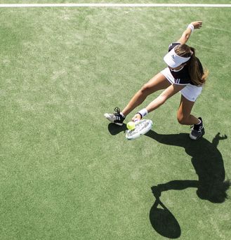 Eine braungebrannte junge Frau in Tenniskleidung befindet sich auf einem Tennisplatz und spielt gerade den Ball zurück, während die Sonne einen schwarzen Schatten auf den grünen Platz wirft.