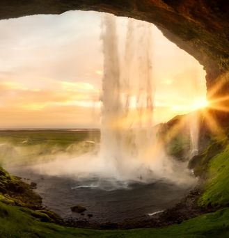 Aus einer Felshöhle heraus führt der Blick in eine weite grüne Landschaft mit untergehender Sonne. Von oben führt ein kräftiger Wasserfall auf die schwarze Erde.