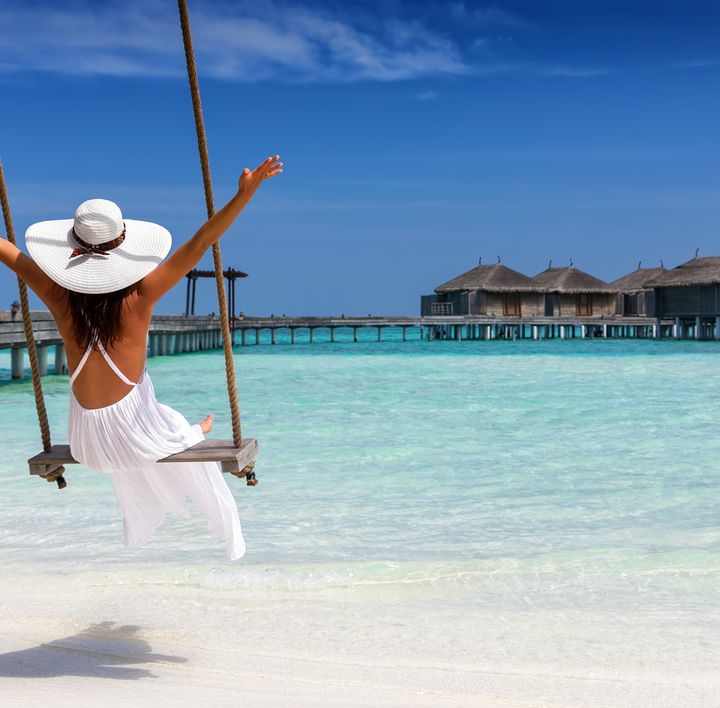 Strandurlaub: Eine Frau mit Sonnenhut und weißem Kleid sitzt auf einer Schaukel an einem türkisen Wasser mit Wasserbungalows