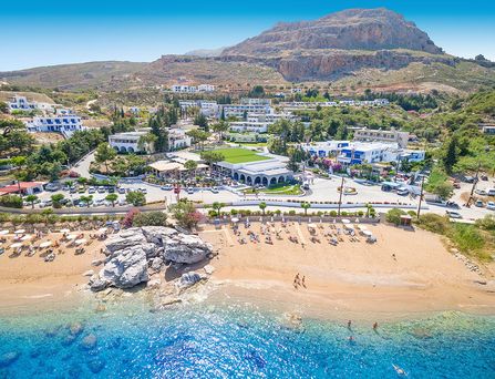 An einem Sandstrand mit einigen Felsen und klarem Wasser stehen viele Sonnenliegen. Dahinter befindet sich eine Hotelanlage auf einem Hang.