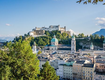 Auf einem Berg befindet sich eine historische Burg und im Tal erstreckt sich die Salzburger Skyline mit vielen hübschen, historischen Gebäuden. Im Hintergrund die Berglandschaft und die Stadt wird immer wieder von Bäumen durchzogen.