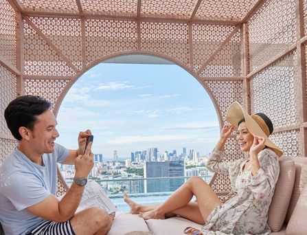 Ein asiatisches Pärchen sitzt in einer modernen Cabana und er schießt ein Foto von ihr. Durch ein rundes Loch ist die Skyline von Singapur zu sehen.