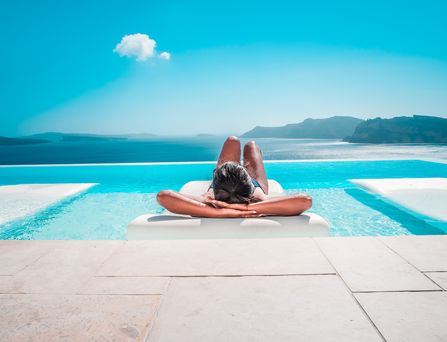 Eine braungebrannte junge Frau in Badekleidung, liegt entspannt auf einer weißen liege auf einem türkisen Infinitypool. Von diesem aus hat man einen unglaublichen Ausblick auf das Meer mit Bergen.