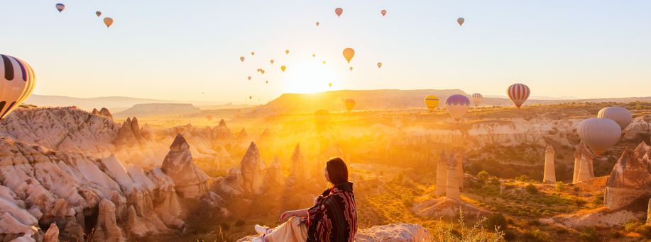 Eine Frau sitzt an einem Felsabsprung und blickt auf die Schlucht von Kappadokien. DIe Sonne geht gerade unter und taucht das gesamte Bild in ein orangenes Licht. Unzählige Heißluftballons schweben in der Luft.